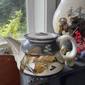 May include: A ceramic teapot with a white handle and spout, and a lid. The teapot is decorated with brown, black, and green abstract designs. A large ceramic vase is in the background.