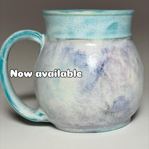 May include: A handcrafted ceramic mug with a rounded form and a substantial handle. The mug has a light blue rim and handle, with a marbled effect of blue, purple, and white on the body. The text "Now available" is visible on the mug.