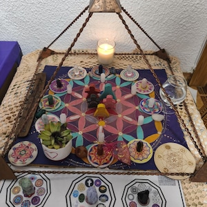 May include: A spiritual display featuring a metal pyramid, a lit candle, and a central board with colourful geometric designs. The arrangement includes small figurines, crystals, and decorative plates, creating a holistic and artistic presentation.