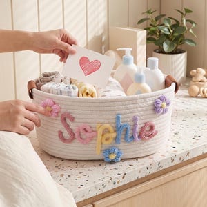 Può includere: Un cesto di corda bianco con il nome "Sophie" in lettere colorate e accenti floreali. Il cesto contiene articoli per bambini ed è tenuto da una persona che indossa un vestito color crema. Una carta con un cuore è tenuta sopra il cesto.
