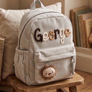 Peut inclure: Un sac à dos en velours côtelé gris clair avec le prénom George brodé en lettres de style corde marron et beige. La poche avant présente un petit écusson rond en forme de tête de lion en peluche avec une crinière marron et une tirette tressée.