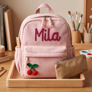 Può includere: Uno zaino in velluto a coste rosa chiaro con il nome Mila ricamato in filo rosa scuro sul davanti. La borsa include una tasca frontale con cerniera decorata con un'applicazione a forma di ciliegie all'uncinetto, un manico superiore e due tasche.