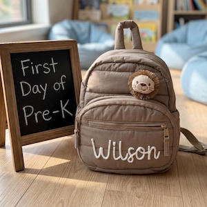 Könnte beinhalten: Ein taupefarbener, gesteppter Rucksack mit einem kleinen Plüsch-Löwengesicht-Aufnäher und dem in weißer Schreibschrift gestickten Namen Wilson. Daneben steht eine hölzerne A-förmige Tafel mit der Aufschrift First Day of Pre-K in weißer Kreide.