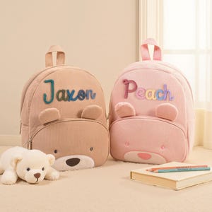 Può includere: Due zaini a forma di orso in velluto a coste, uno beige e uno rosa, con i nomi ricamati "Jaxon" e "Peach". Gli zaini hanno orecchie e facce da orso. Un orsacchiotto bianco è davanti allo zaino beige.
