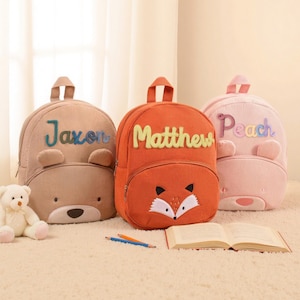 Könnte beinhalten: Drei Rucksäcke aus Cord in den Farben Beige, Orange und Rosa, jeweils mit einem anderen Tiergesicht-Design. Der beige Rucksack hat "Jaxon" in Blau gestickt, der orangefarbene Rucksack hat ein Fuchgesicht und "Matthew" in Gelb, und der rosa Rucksack hat "Peach" in Rosa.