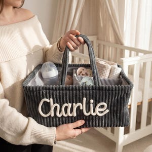 Op de afbeelding: Een grijze babycaddy van corduroy met de naam "Charlie" in witte letters. De caddy bevat een babyfles, fopspenen en kleine dekentjes. De caddy heeft een gestreepte binnenkant en een handvat.