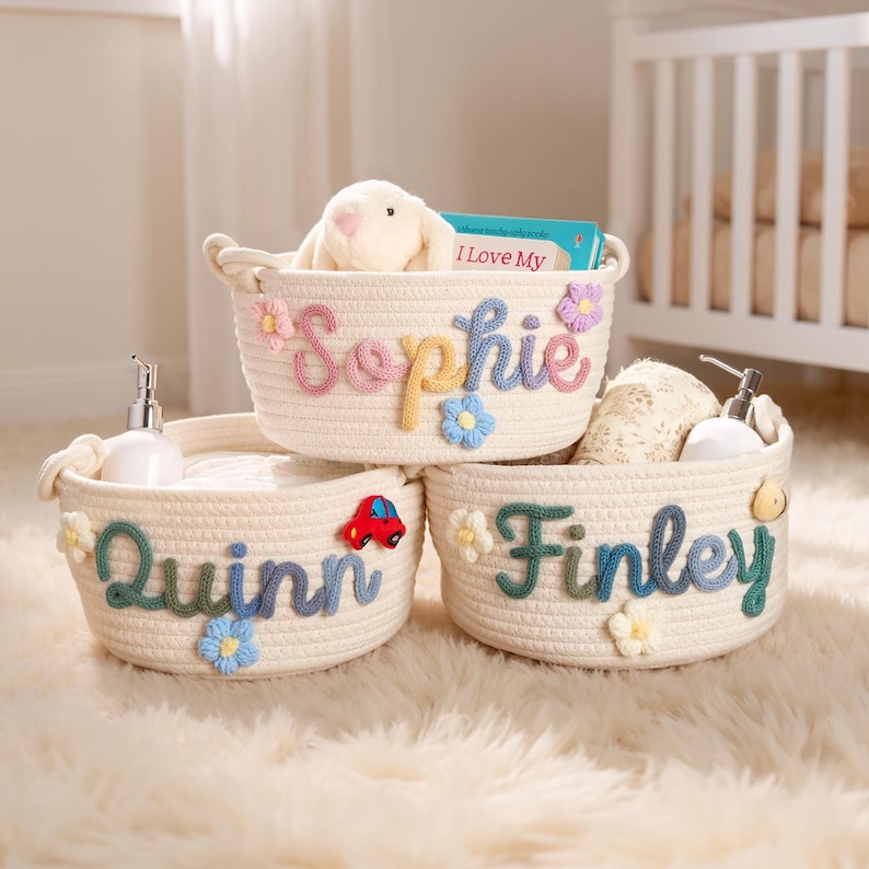 May include: Three cream-colored rope baskets with embroidered names: "Sophie," "Quinn," and "Finley." Each basket has floral and car embellishments. The "Sophie" basket contains a stuffed bunny and a book. The baskets sit on a fluffy white rug.
