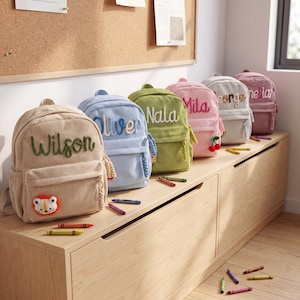 Könnte beinhalten: Eine Reihe von sechs Rucksäcken aus Kord in Beige, Hellblau, Olivgrün, Rosa, Hellgrau und Mauve. Jeder Rucksack ist mit einem aufgestickten Namen auf der Vorderseite und einem kleinen Plüschtier- oder Fruchtanhänger am Reißverschluss versehen.