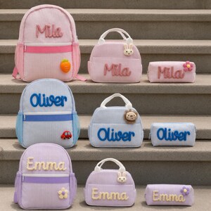 Op de afbeelding: Een collectie van drie sets corduroy tassen, elk bestaande uit een rugzak, een lunchtas en een etui. De sets zijn gepersonaliseerd met de namen Mila in roze, Oliver in blauw en Emma in paars, voorzien van kleine pluche iconen.