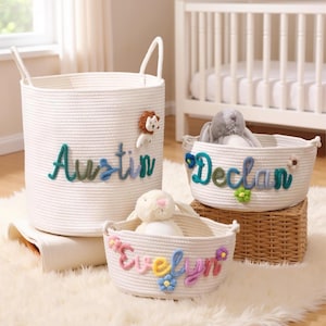May include: Three white rope baskets with personalized names: "Austin", "Declan", and "Evelyn". The "Austin" basket is cylindrical with handles and a lion applique. The other two are oval-shaped, with floral accents and stuffed animals inside.
