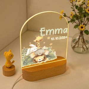 Könnte beinhalten: Personalisiertes Nachtlicht mit dem Namen "Emma" und dem Datum "10.10.2024" auf einer klaren, gewölbten Platte beleuchtet. Die Platte zeigt eine farbenfrohe Illustration einer Giraffe, eines Elefanten, eines Löwen und eines Kaninchens. Der Sockel ist aus Holz.