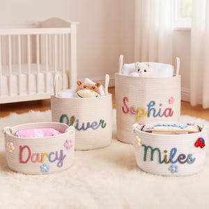 May include: Four cream-colored rope storage baskets with handles, each with a child's name in colorful letters. The baskets are filled with toys and blankets. The names are Darcy, Oliver, Sophia, and Miles. Decorative flowers and a car are attached to the baskets.