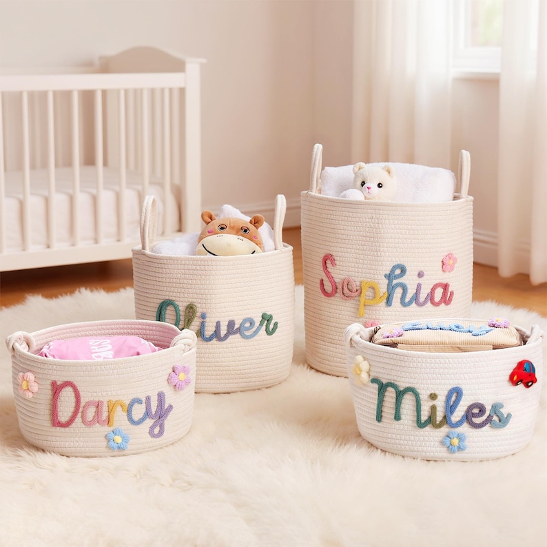 May include: Four cream-colored rope storage baskets with handles, each with a child's name in colorful letters. The baskets are filled with toys and blankets. The names are Darcy, Oliver, Sophia, and Miles. Decorative flowers and a car are attached to the baskets.