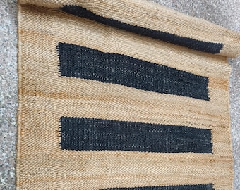 Puede incluir: Una alfombra rectangular de yute con tres franjas horizontales negras. La alfombra está parcialmente enrollada, mostrando su textura de fibra natural y acentos oscuros contrastantes. Adecuada para la decoración del hogar.