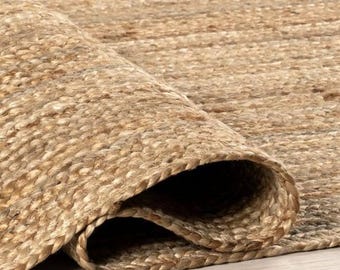 Puede incluir: Primer plano de una alfombra de yute enrollada, en tonos naturales. La textura de la alfombra es visible, con fibras densamente tejidas que crean una superficie duradera. El borde enrollado revela el grosor y la construcción de la alfombra, adecuada para la decoración del hogar.