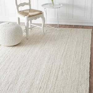 May include: A cream-coloured, textured rug with a woven pattern. The rug is rectangular and covers a wooden floor. A white chair, a small side table, and a round pouf are in the background.