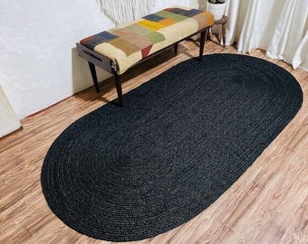 May include: An oval-shaped area rug crafted from braided black jute fibers. The rug features a continuous, concentric coil pattern that creates a textured, uniform surface across the entire piece, providing a solid dark foundation.