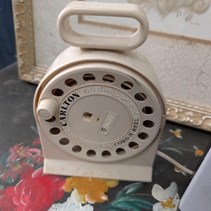 May include: A beige plastic Carlton Cord-N-Reel device featuring a circular housing with a series of ventilation holes, a central winding knob, and a top-mounted carrying handle. The brand name and product name are printed on the front face.