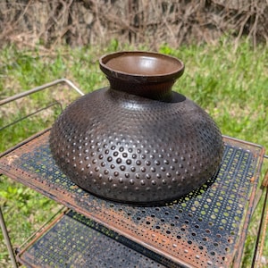 May include: A dark brown, textured vase with a rounded body and a small, flared opening. The surface is covered in small, evenly spaced bumps. The vase sits on a rusty, metal plant stand with multiple shelves, outdoors.