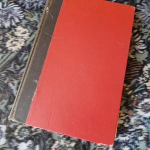 May include: A hardback book featuring a bright red front cover and a dark grey spine. The book has a rectangular shape with visible wear along the edges of the spine and cover, showing a textured surface and a simple, solid-coloured design.