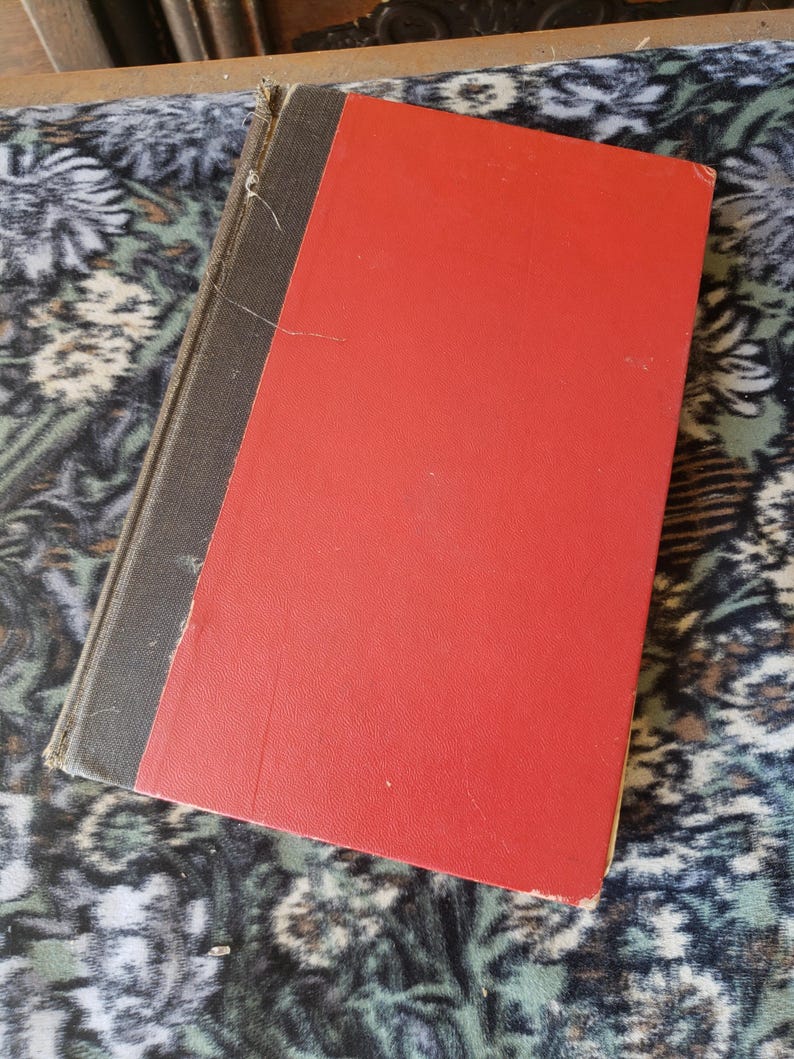 May include: A hardback book featuring a bright red front cover and a dark grey spine. The book has a rectangular shape with visible wear along the edges of the spine and cover, showing a textured surface and a simple, solid-coloured design.