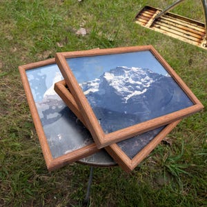 May include: Two rectangular picture frames with light brown wooden borders, each containing a photographic print of a snow-capped mountain peak under a clear blue sky. The frames are stacked at an angle, showcasing the mountain imagery.