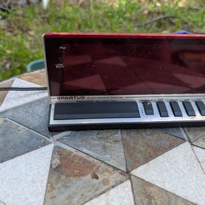 Könnte beinhalten: Ein Vintage Spartus elektronischer Digitalwecker mit roter Anzeige. Die Uhr hat ein silbernes und schwarzes Bedienfeld im Tastaturstil mit Tasten für Zeit-, Stunden- und Minuteneinstellungen. Das Display zeigt "PM" und "LOW BATT".