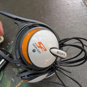 May include: Sony MDR-G57 headphones featuring a white circular ear cup with an orange textured accent ring. The device includes a black plastic frame, a black cord, and the Sony logo printed in black on a white tab extending from the side.