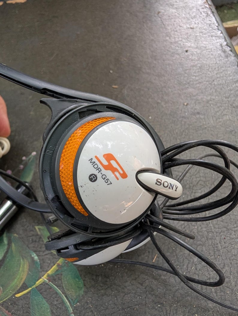 May include: Sony MDR-G57 headphones featuring a white circular ear cup with an orange textured accent ring. The device includes a black plastic frame, a black cord, and the Sony logo printed in black on a white tab extending from the side.