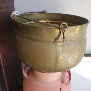 May include: A large, round brass basin with a weathered, golden-toned surface. It features a curved metal handle attached to two side loops, with a small, cylindrical white ceramic grip decorated with a blue floral pattern on one side.