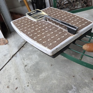 Puede incluir: Báscula de baño vintage con un patrón marrón y blanco, asa cromada y base negra. La báscula tiene un borde blanco y una cubierta de esfera de plástico transparente. Se encuentra sobre una estantería metálica verde.