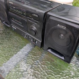 Puede incluir: Un sistema estéreo portátil Sony CFS-1025 de color negro que incluye un reproductor de casetes, un sintonizador de radio analógico con escala de frecuencia, una perilla de volumen y dos altavoces desmontables con rejillas metálicas.
