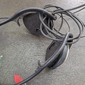 May include: Black wired headphones featuring a thin, flexible headband and two circular ear cups with black foam padding. The thin black audio cable is coiled and tangled around the headband structure, connecting to the circular ear pieces.