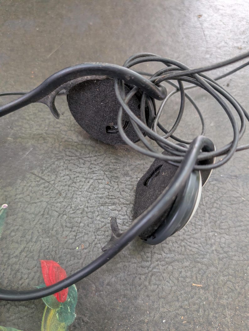 May include: Black wired headphones featuring a thin, flexible headband and two circular ear cups with black foam padding. The thin black audio cable is coiled and tangled around the headband structure, connecting to the circular ear pieces.