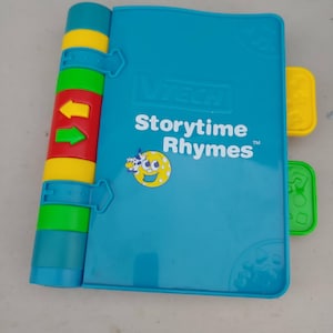 May include: A VTech Storytime Rhymes electronic book toy with a teal cover, featuring a yellow smiling moon graphic and colourful plastic buttons along the spine and side. The front cover displays the text VTech Storytime Rhymes in white letters.