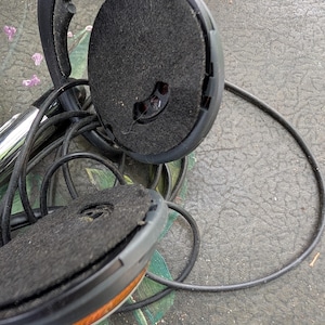 May include: A pair of black headphones with circular ear cups featuring dark grey felt padding and a thin black cord. The plastic frame is visible, showing a hinge mechanism and a small opening in the center of the felt ear cushion.
