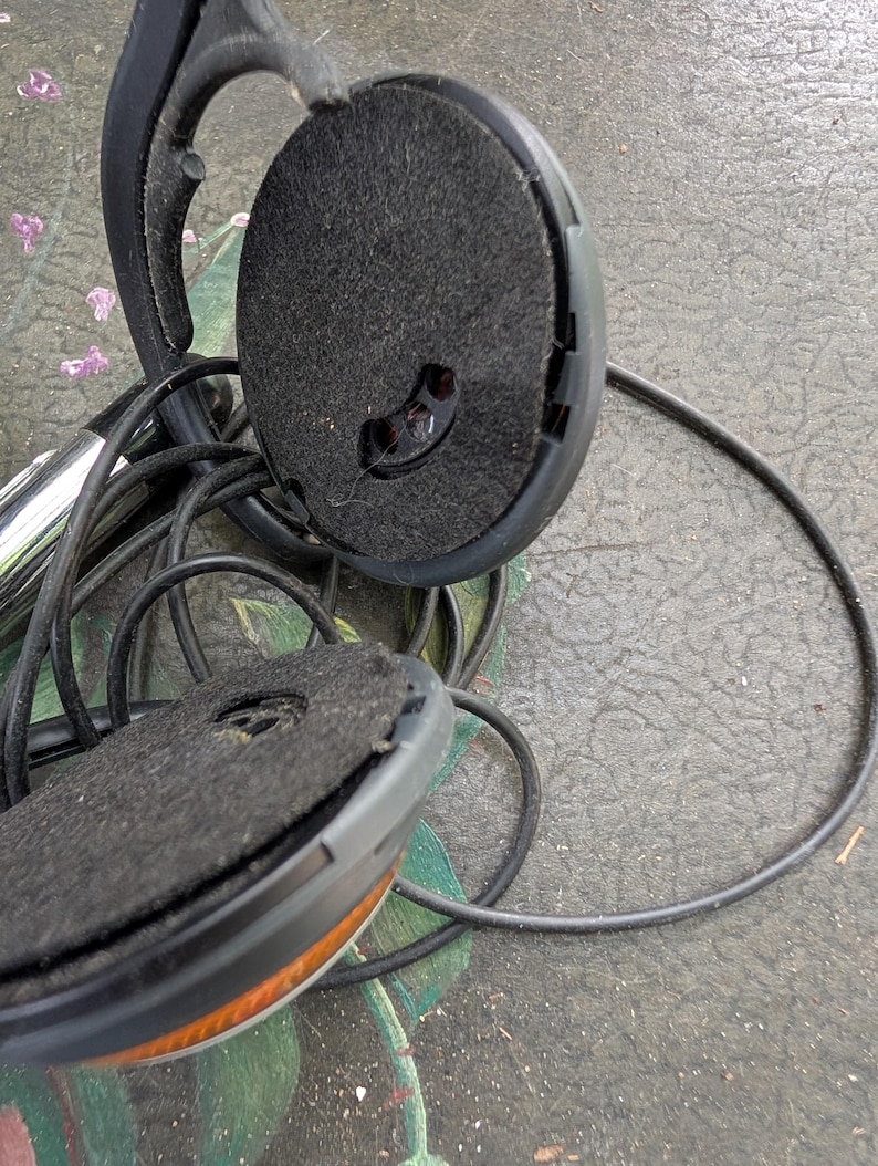 May include: A pair of black headphones with circular ear cups featuring dark grey felt padding and a thin black cord. The plastic frame is visible, showing a hinge mechanism and a small opening in the center of the felt ear cushion.