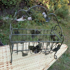 May include: A rectangular wire basket with a dark green finish, featuring a decorative arched handle and metal leaf and flower accents attached to the sides. The basket stands on four small curved feet and has an open grid structure.