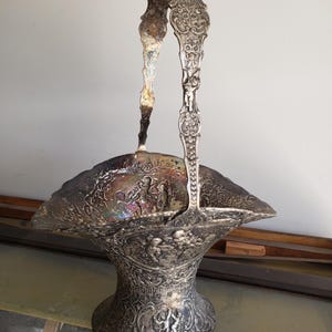May include: A silver-toned metal basket featuring an ornate, raised relief design of cherubs and intricate floral scrollwork. The piece includes a tall, arched handle and a wide, flared rim with a tarnished, iridescent patina finish.