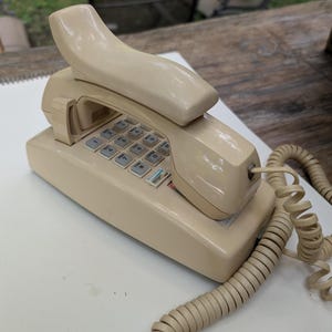 May include: A beige landline telephone with a rectangular base and a matching handset resting on top. The device features a standard numeric keypad with twelve buttons, including an asterisk and a pound sign, and a coiled cord extending from the side.