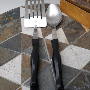 May include: A stainless steel spatula and spoon with black handles rest on a patterned surface. The spatula has a slotted design, and the spoon has a rounded bowl. The handles are a glossy black, and the set appears to be kitchen utensils.