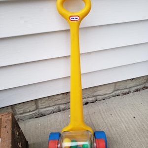 May include: A yellow Little Tikes push toy with a handle and a clear plastic cylinder containing colorful balls. The wheels are red and blue. The toy is designed for toddlers and young children.
