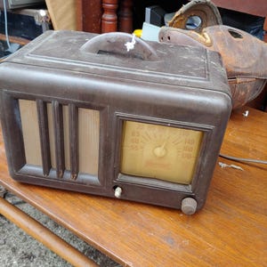 Op de afbeelding: Een vintage bruine General Electric tafelradio met een rechthoekige behuizing, een handvat aan de bovenzijde, een luidsprekerrooster met drie verticale sleuven en een geel getint wijzerplaatvenster met frequentiegetallen van 55 tot 170.