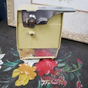 May include: A vintage Rival electric can opener and knife sharpener with a pale yellow textured plastic body and a chrome metal top mechanism. The front panel features the Rival logo and the text can opener / knife sharpener in black.