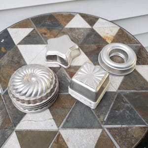 Puede incluir: Una colección de moldes para hornear de metal vintage en varias formas, incluyendo una estrella, un cuadrado, un anillo y un molde redondo estriado. Los moldes son plateados y parecen ser de aluminio. Los moldes están colocados sobre una mesa de mosaico.