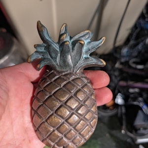 May include: A pineapple-shaped decorative object made of metal with a bronze-toned finish on the body and a dark, oxidized green patina on the leafy top. The surface features a textured, diamond-patterned grid across the fruit section.