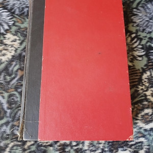 May include: A hardback book featuring a bright red front cover and a dark grey spine. The book has a rectangular shape with visible wear along the edges of the spine and corners of the cover, showing signs of age and use on the binding material.