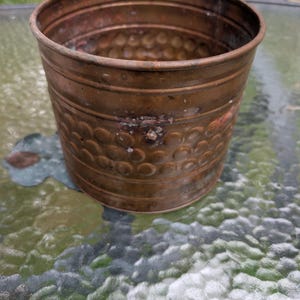 Pode incluir: Um vaso cilíndrico de cobre com um exterior texturizado apresentando um padrão repetitivo de pequenas reentrâncias circulares. A superfície metálica exibe uma pátina envelhecida com manchas escuras e oxidação visível por todo o corpo.