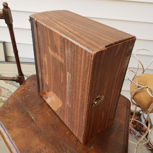 May include: A rectangular wooden box with a hinged lid and a brass clasp. The box has a dark brown finish with visible wood grain and some areas of wear. It sits on a wooden table, suggesting a vintage or antique item.