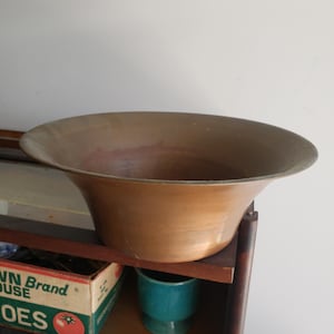 May include: A wide-rimmed, metallic bowl with a warm, brownish-copper tone and a smooth, curved exterior. The bowl features a deep, open basin with a slightly darkened interior surface, showcasing a simple, functional design with a flared edge.